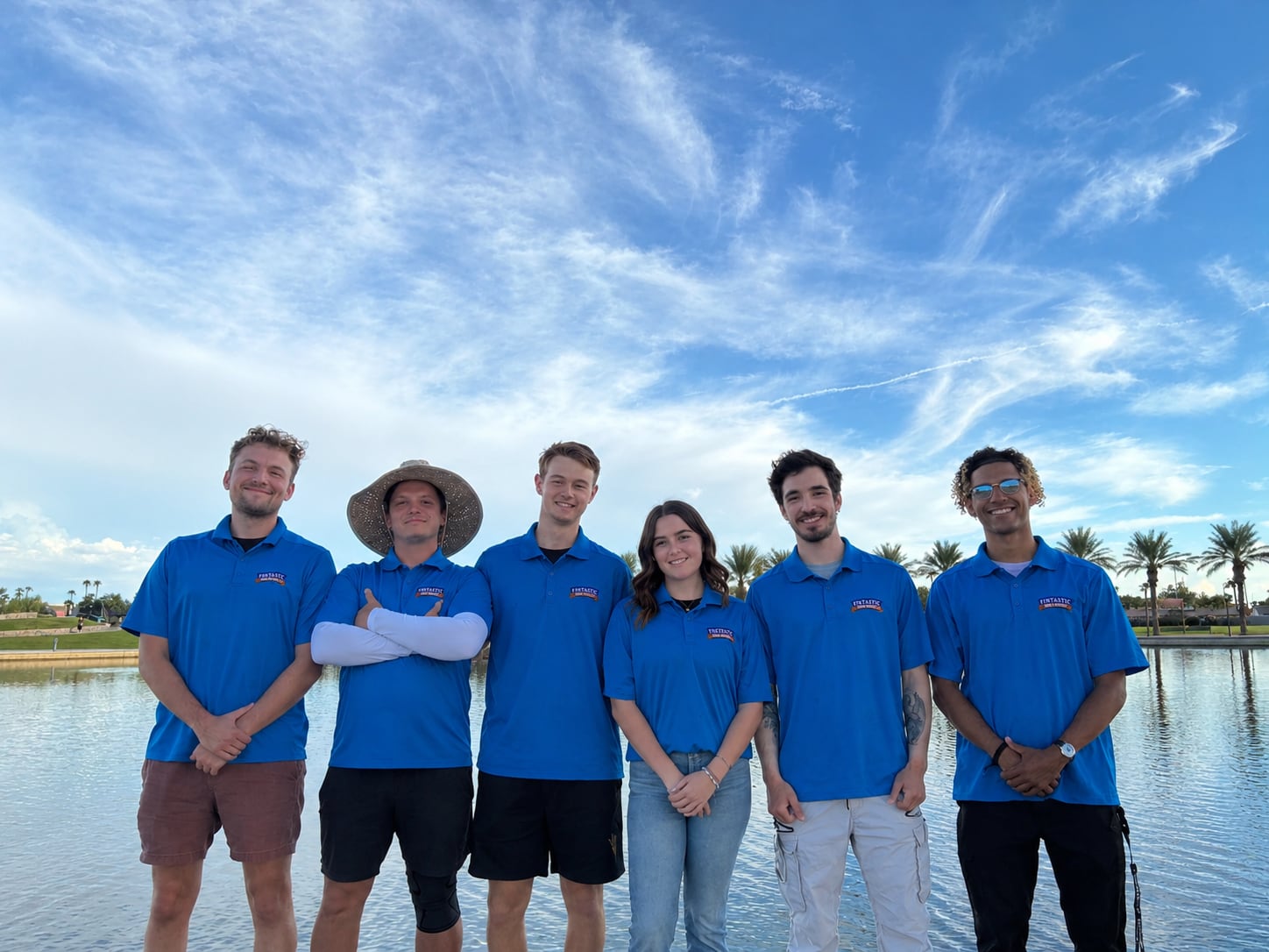 Finn’s pool service team — six people in matching blue shirts standing at the edge of a lake under a bright sky.